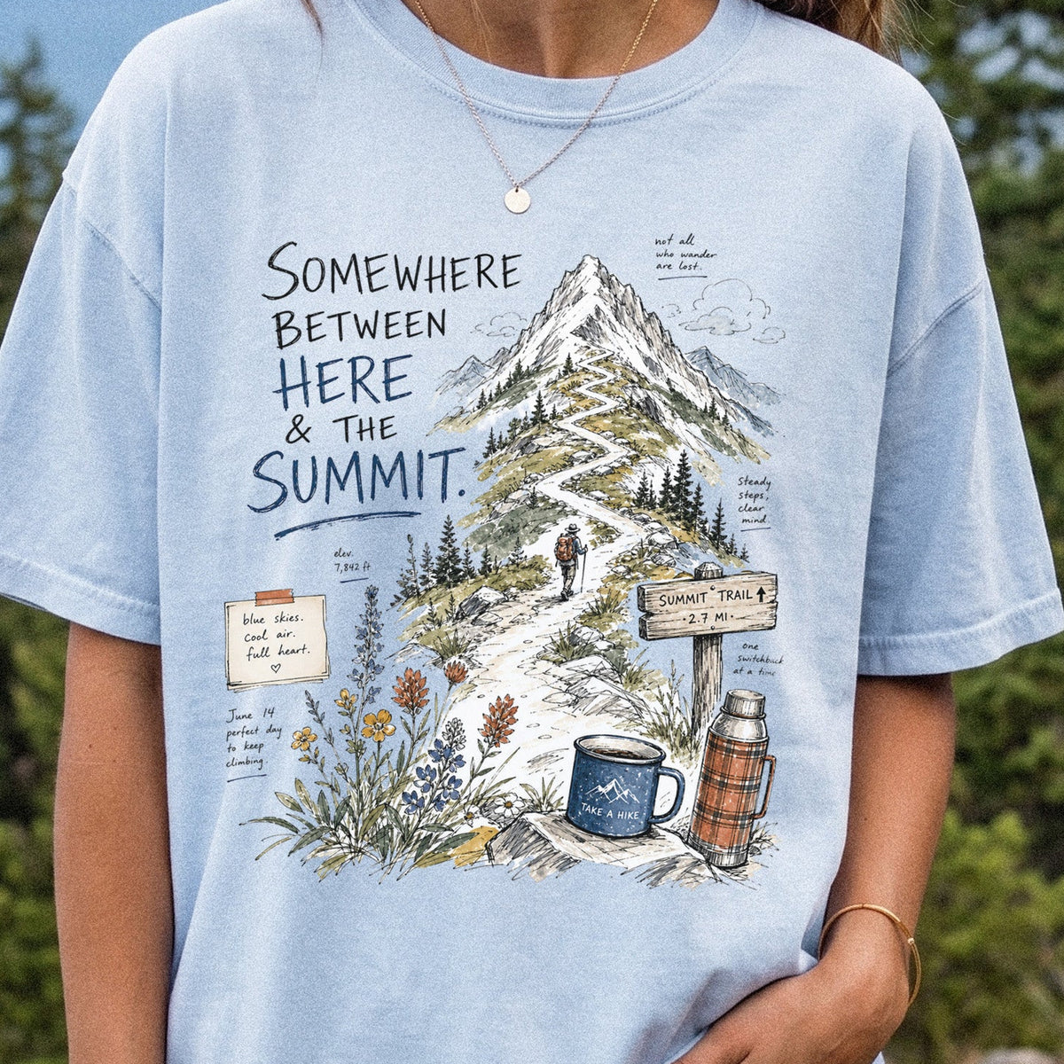 Woman wearing a Chambray T-shirt printed with a hand-drawn mountain summit trail scene with wildflowers, a trail sign, a hiker, and a camp mug