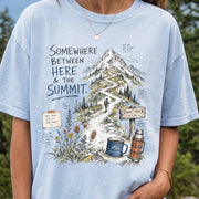 Woman wearing a Chambray T-shirt printed with a hand-drawn mountain summit trail scene with wildflowers, a trail sign, a hiker, and a camp mug