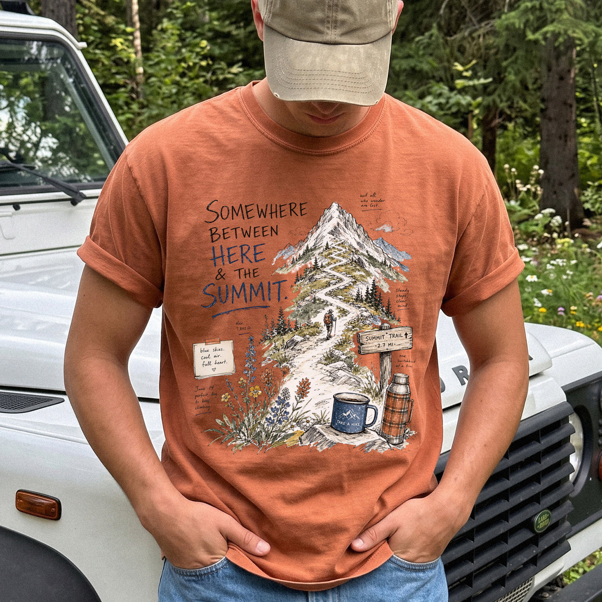 Man wearing a Yam T-shirt printed with a hand-drawn mountain summit trail scene with wildflowers, a trail sign, a hiker, and a camp mug
