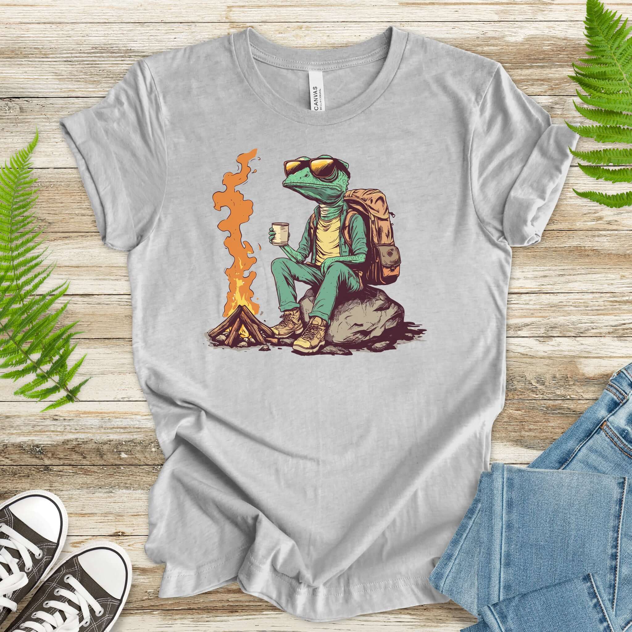 Chameleon in hiking gear sitting near a campfire, wearing sunglasses and holding a drink on a gray t-shirt.