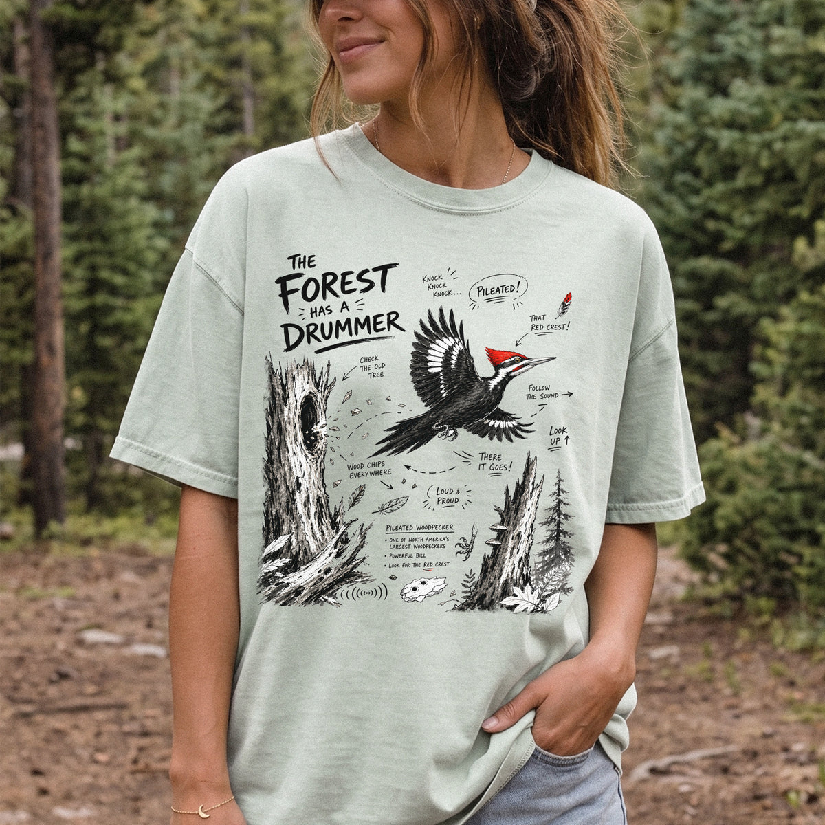The Forest Has a Drummer Comfort Colors Shirt – Pileated Woodpecker design of a Pileated Woodpecker illustrated in flight with its vivid red crest, surrounded by old tree trunks, wood chips, and forest details, printed on a Bay T-shirt, worn by a woman.