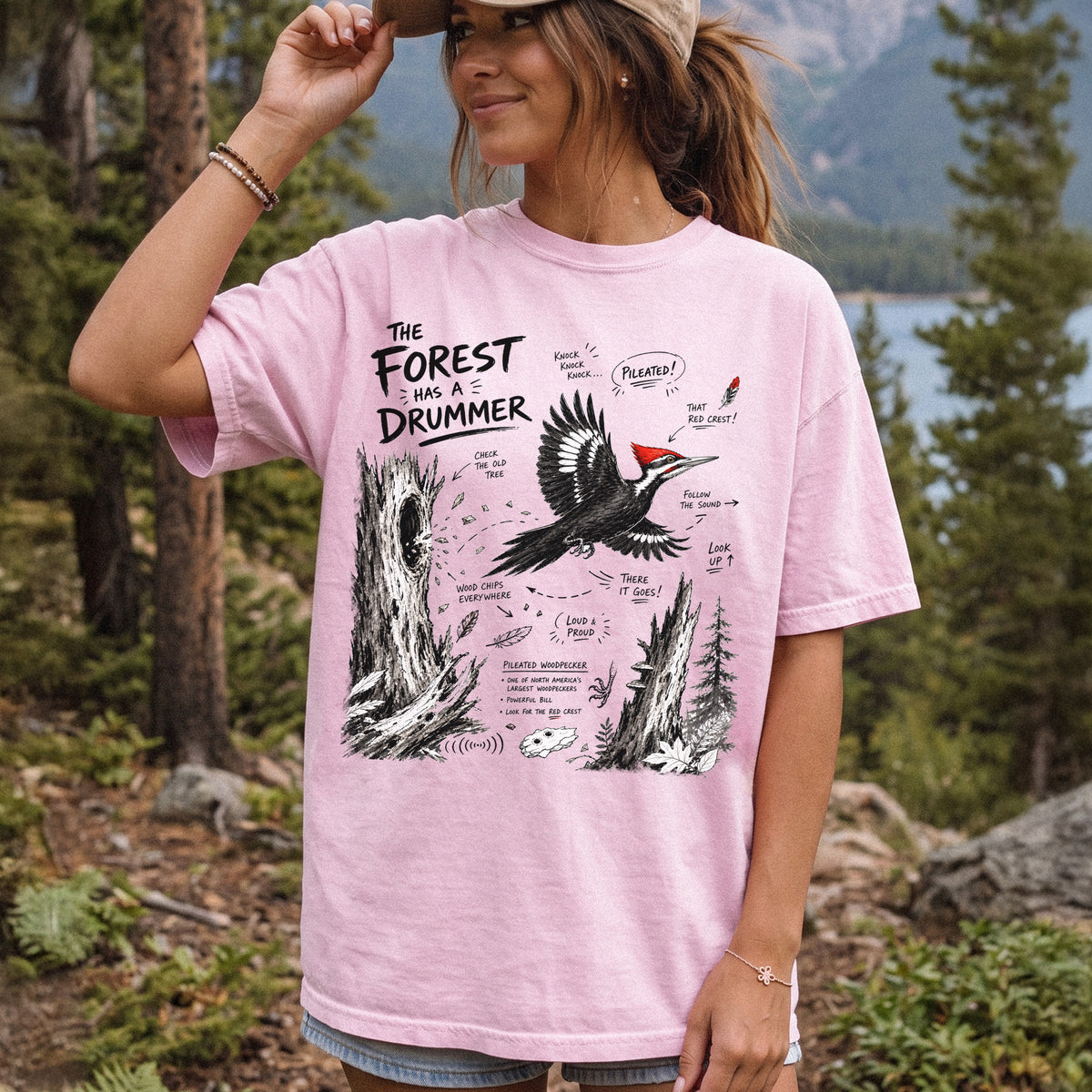 The Forest Has a Drummer Comfort Colors Shirt – Pileated Woodpecker design of a Pileated Woodpecker illustrated in flight with its vivid red crest, surrounded by old tree trunks, wood chips, and forest details, printed on a Blossom T-shirt, worn by a woman.