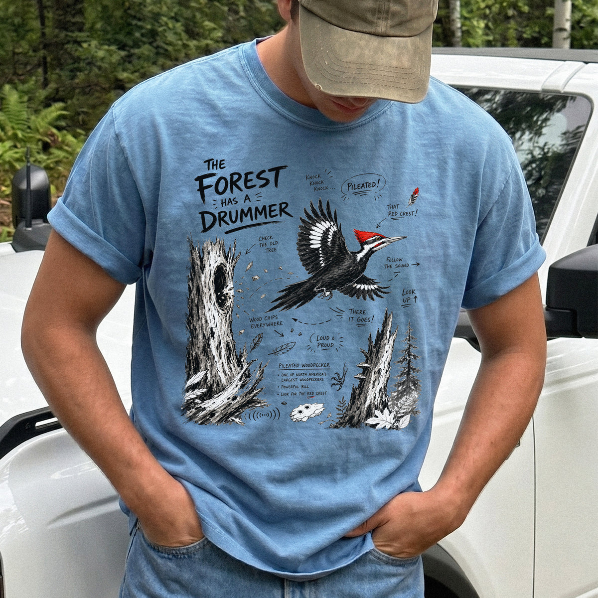 The Forest Has a Drummer Comfort Colors Shirt – Pileated Woodpecker design of a Pileated Woodpecker illustrated in flight with its vivid red crest, surrounded by old tree trunks, wood chips, and forest details, printed on a Blue Jean T-shirt.