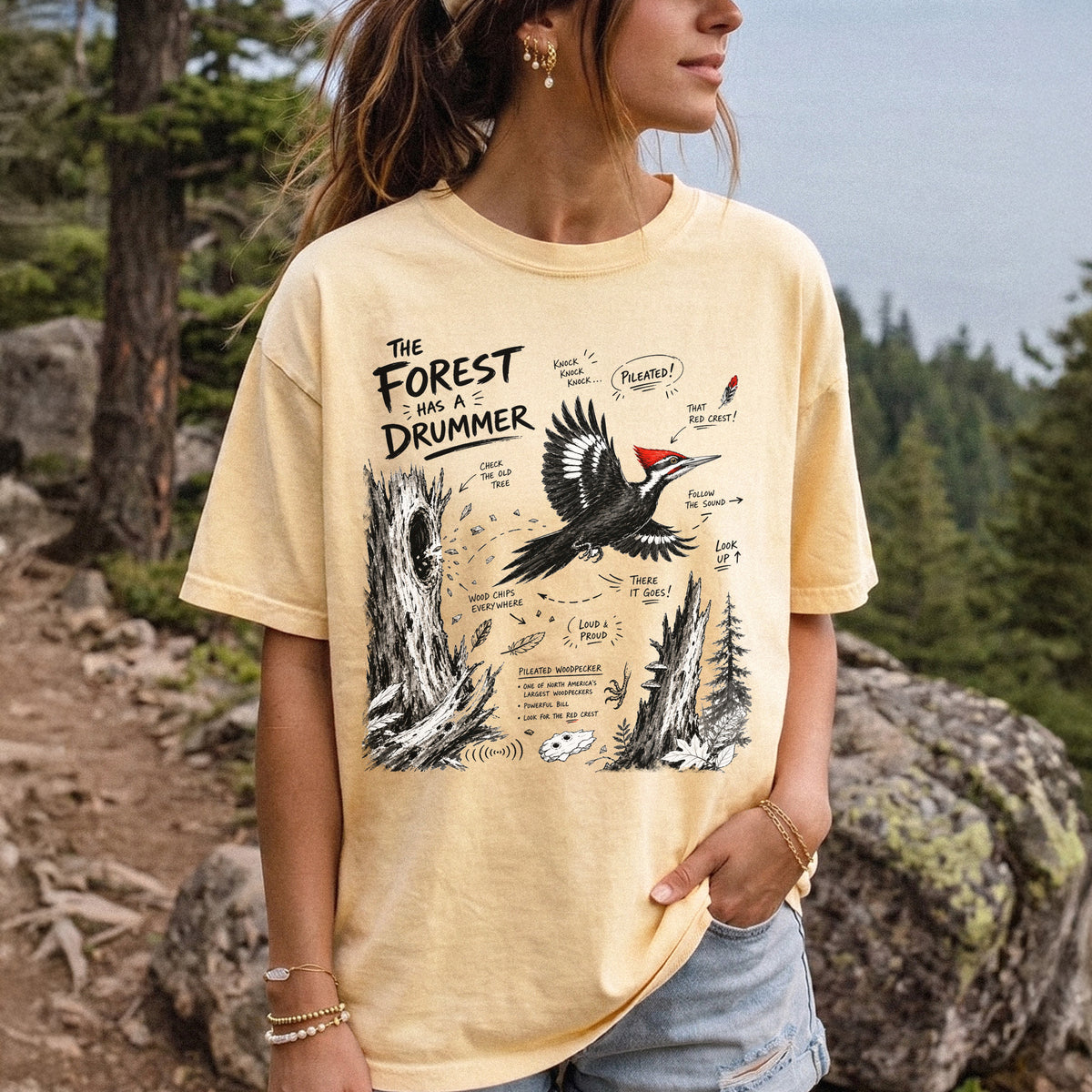The Forest Has a Drummer Comfort Colors Shirt – Pileated Woodpecker design of a Pileated Woodpecker illustrated in flight with its vivid red crest, surrounded by old tree trunks, wood chips, and forest details, printed on a Butter T-shirt, worn by a woman.