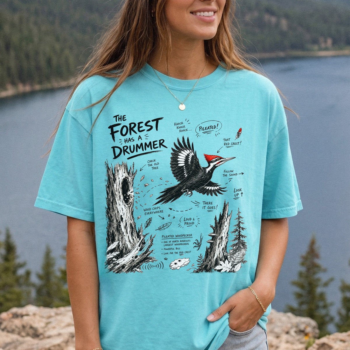The Forest Has a Drummer Comfort Colors Shirt – Pileated Woodpecker design of a Pileated Woodpecker illustrated in flight with its vivid red crest, surrounded by old tree trunks, wood chips, and forest details, printed on a Chalky Mint T-shirt, worn by a woman.