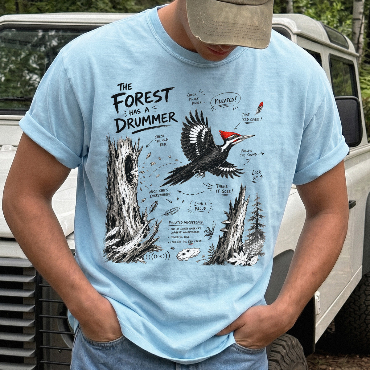 The Forest Has a Drummer Comfort Colors Shirt – Pileated Woodpecker design of a Pileated Woodpecker illustrated in flight with its vivid red crest, surrounded by old tree trunks, wood chips, and forest details, printed on a Chambray T-shirt.