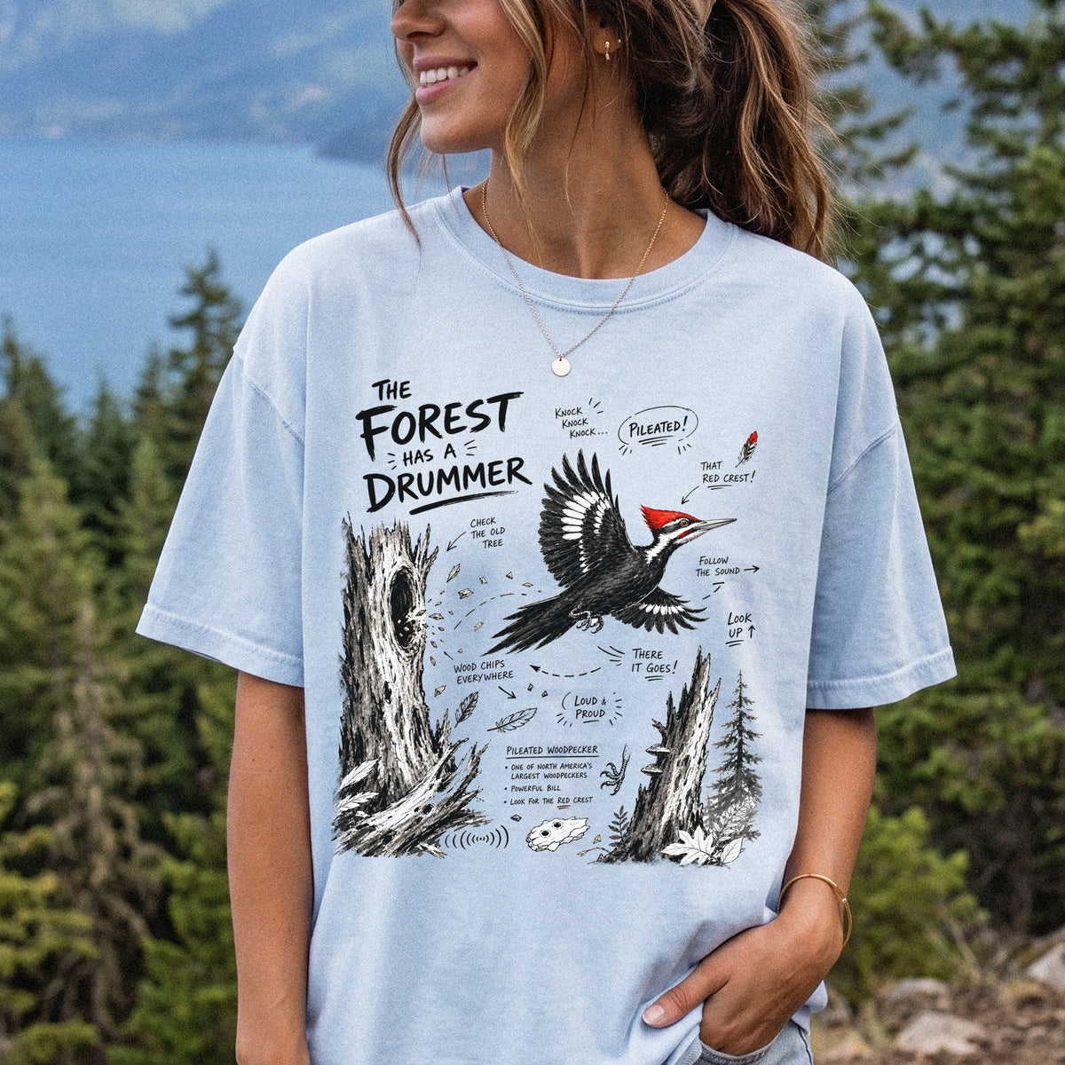 The Forest Has a Drummer Comfort Colors Shirt – Pileated Woodpecker design of a Pileated Woodpecker illustrated in flight with its vivid red crest, surrounded by old tree trunks, wood chips, and forest details, printed on a Chambray T-shirt, worn by a woman.