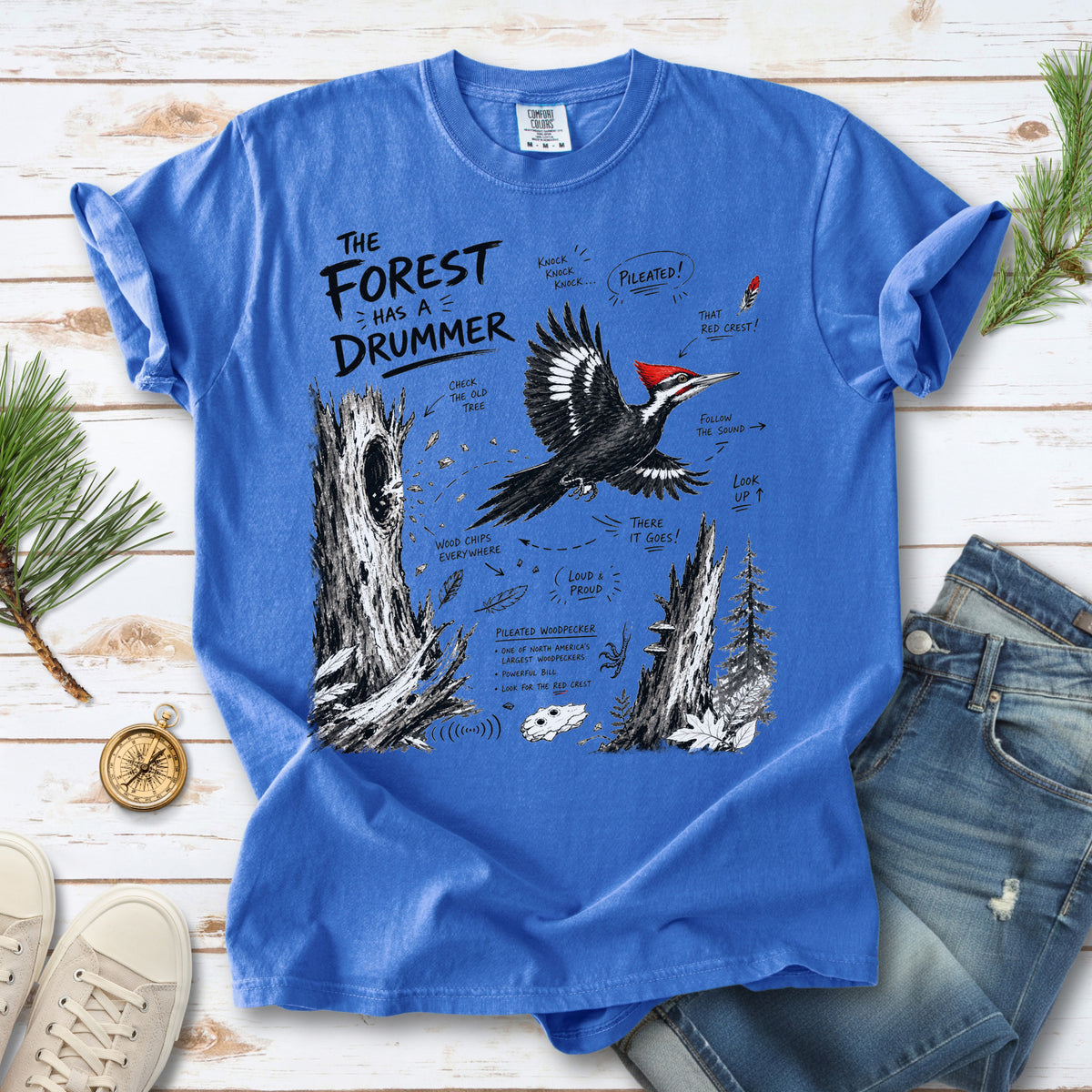 The Forest Has a Drummer Comfort Colors Shirt – Pileated Woodpecker design of a Pileated Woodpecker illustrated in flight with its vivid red crest, surrounded by old tree trunks, wood chips, and forest details, printed on a Flo Blue T-shirt.