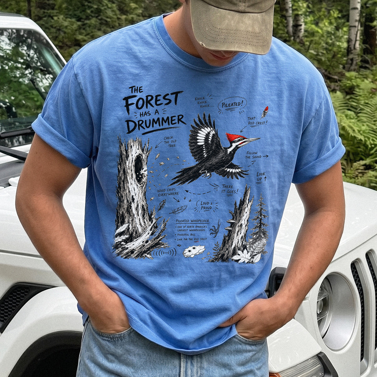 The Forest Has a Drummer Comfort Colors Shirt – Pileated Woodpecker design of a Pileated Woodpecker illustrated in flight with its vivid red crest, surrounded by old tree trunks, wood chips, and forest details, printed on a Flo Blue T-shirt.