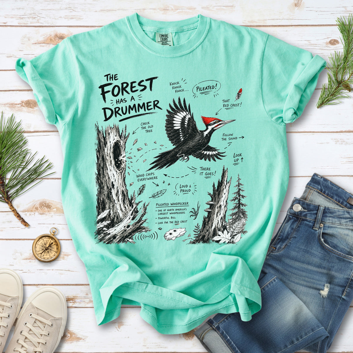 The Forest Has a Drummer Comfort Colors Shirt – Pileated Woodpecker design of a Pileated Woodpecker illustrated in flight with its vivid red crest, surrounded by old tree trunks, wood chips, and forest details, printed on an Island Reef T-shirt.