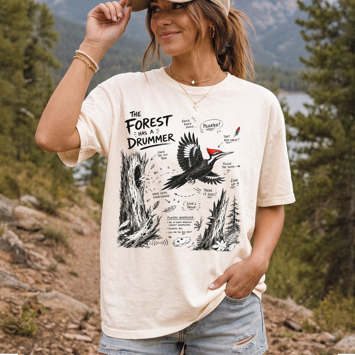 The Forest Has a Drummer Comfort Colors Shirt – Pileated Woodpecker design of a Pileated Woodpecker illustrated in flight with its vivid red crest, surrounded by old tree trunks, wood chips, and forest details, printed on an Ivory T-shirt, worn by a woman.