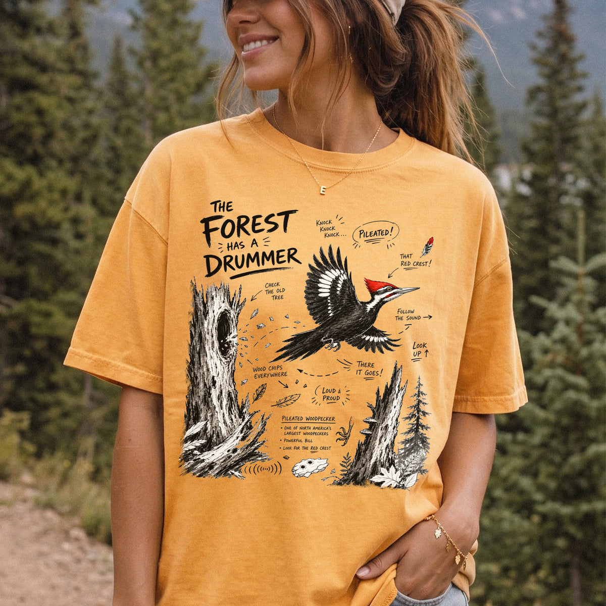 The Forest Has a Drummer Comfort Colors Shirt – Pileated Woodpecker design of a Pileated Woodpecker illustrated in flight with its vivid red crest, surrounded by old tree trunks, wood chips, and forest details, printed on a Mustard T-shirt, worn by a woman.