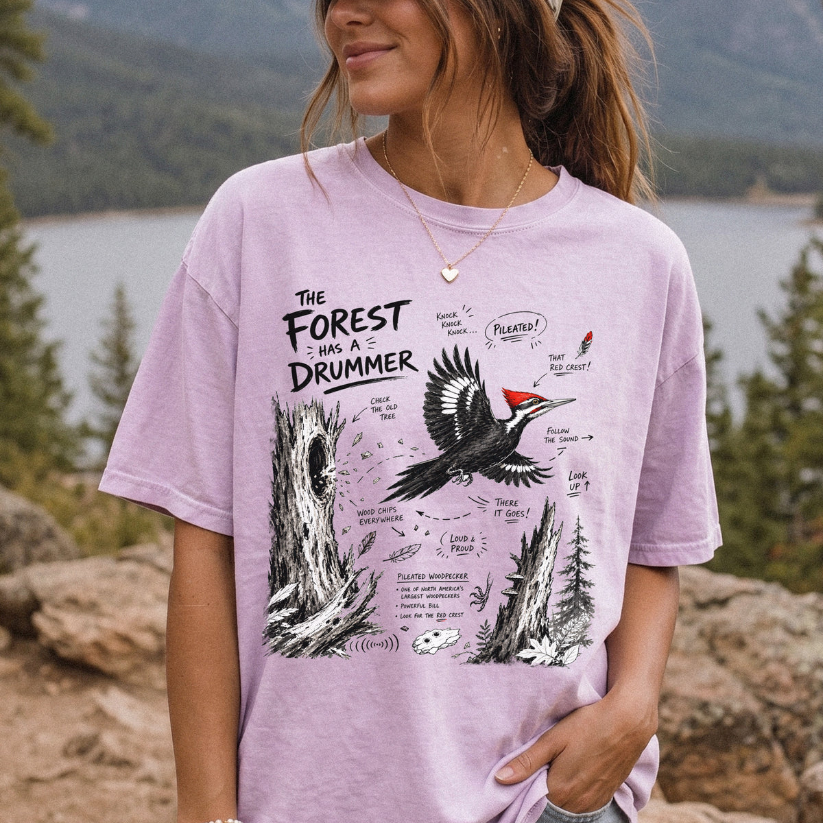 The Forest Has a Drummer Comfort Colors Shirt – Pileated Woodpecker design of a Pileated Woodpecker illustrated in flight with its vivid red crest, surrounded by old tree trunks, wood chips, and forest details, printed on an Orchid T-shirt, worn by a woman.