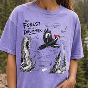 The Forest Has a Drummer Comfort Colors Shirt – Pileated Woodpecker design of a Pileated Woodpecker illustrated in flight with its vivid red crest, surrounded by old tree trunks, wood chips, and forest details, printed on a Violet T-shirt, worn by a woman.