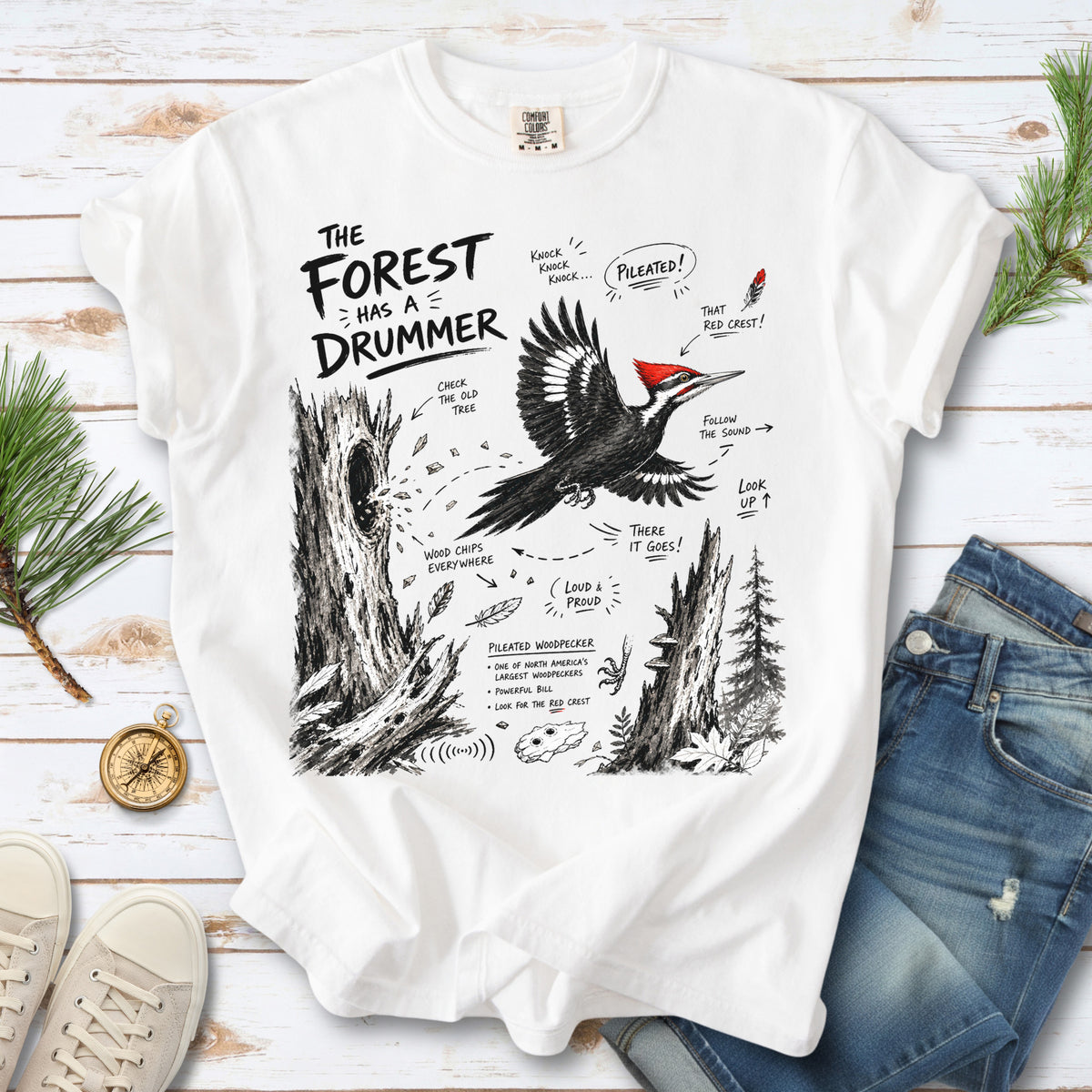 The Forest Has a Drummer Comfort Colors Shirt – Pileated Woodpecker design of a Pileated Woodpecker illustrated in flight with its vivid red crest, surrounded by old tree trunks, wood chips, and forest details, printed on a White T-shirt.