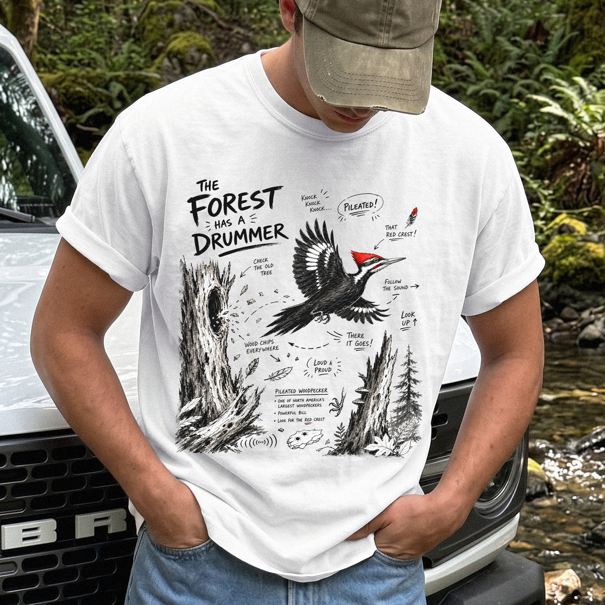 The Forest Has a Drummer Comfort Colors Shirt – Pileated Woodpecker design of a Pileated Woodpecker illustrated in flight with its vivid red crest, surrounded by old tree trunks, wood chips, and forest details, printed on a White T-shirt.