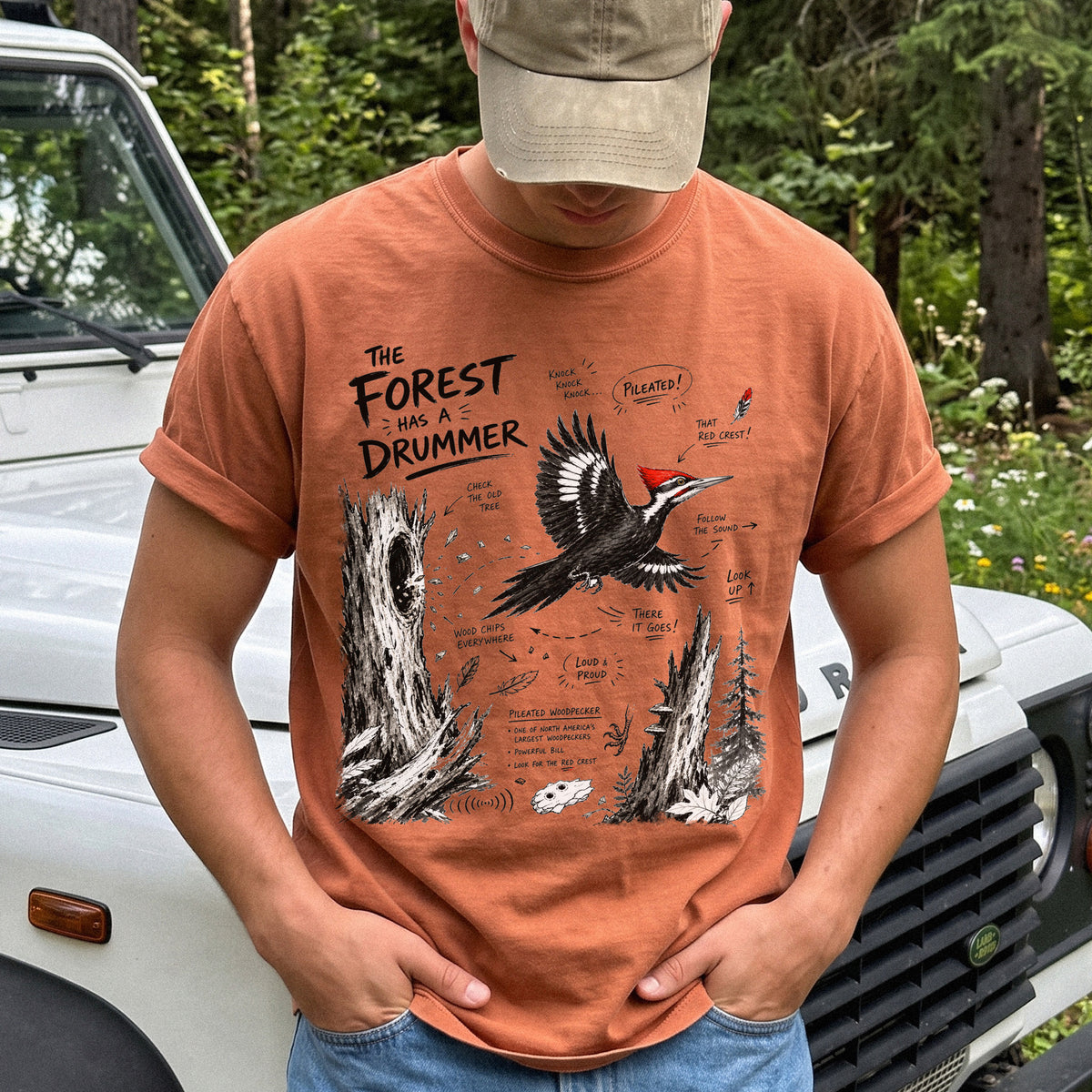 The Forest Has a Drummer Comfort Colors Shirt – Pileated Woodpecker design of a Pileated Woodpecker illustrated in flight with its vivid red crest, surrounded by old tree trunks, wood chips, and forest details, printed on a Yam T-shirt.