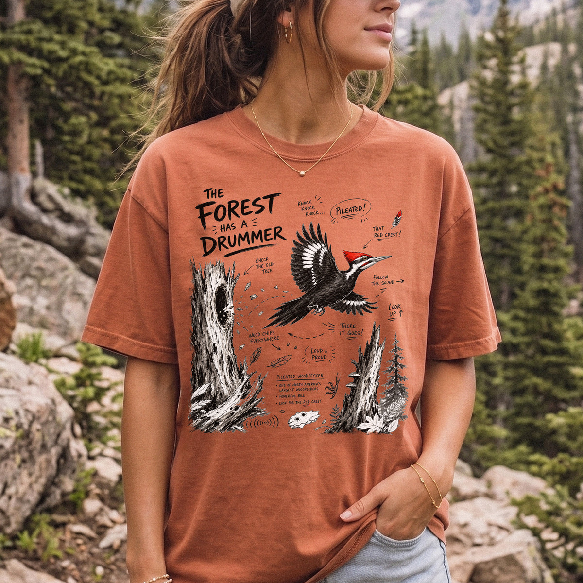 The Forest Has a Drummer Comfort Colors Shirt – Pileated Woodpecker design of a Pileated Woodpecker illustrated in flight with its vivid red crest, surrounded by old tree trunks, wood chips, and forest details, printed on a Yam T-shirt, worn by a woman.