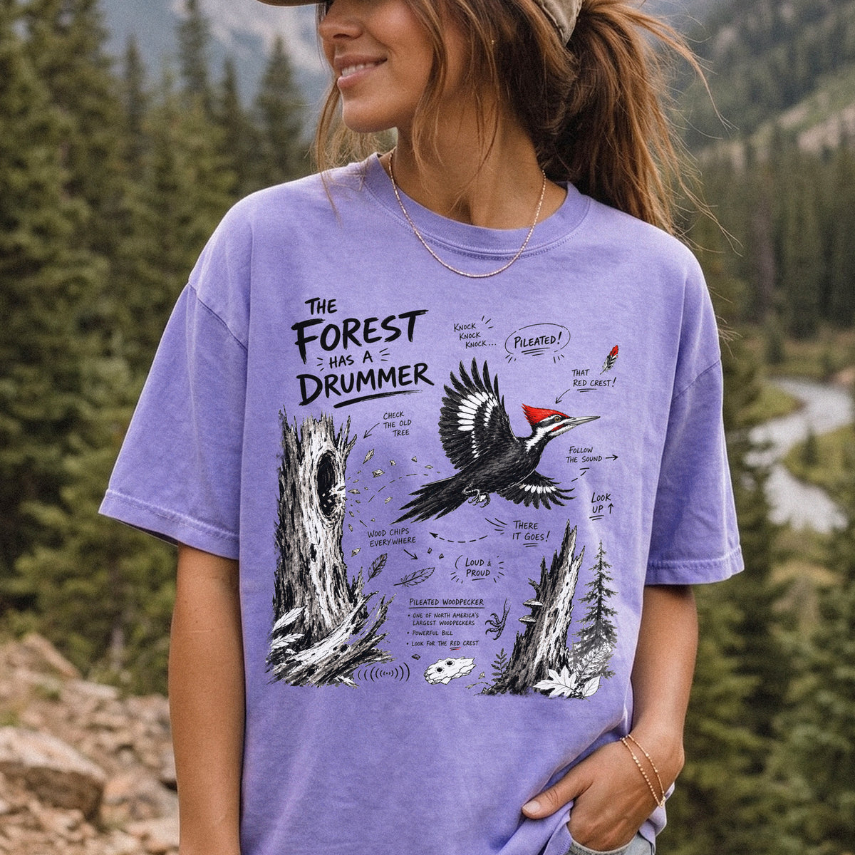 The Forest Has a Drummer Comfort Colors Shirt – Pileated Woodpecker design of a Pileated Woodpecker illustrated in flight with its vivid red crest, surrounded by old tree trunks, wood chips, and forest details, printed on a Violet T-shirt, worn by a woman.