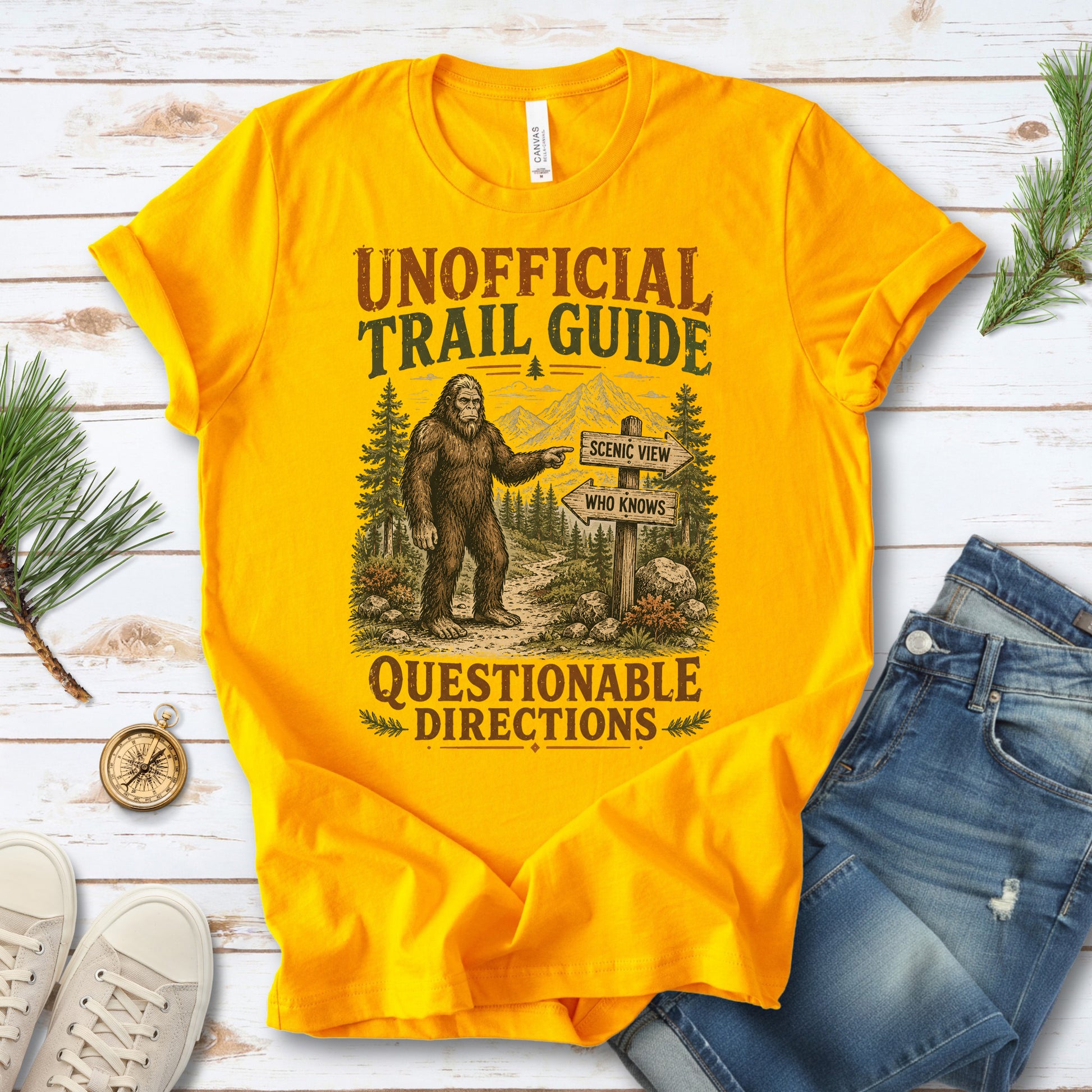 A bright yellow graphic t-shirt laid flat on a white wood background, styled with blue jeans, sneakers, pine branches, and a compass. The t-shirt features a vintage-style graphic of Bigfoot pointing toward a trail sign that reads 'ESCAPE VIEW WHO KNOWS'.