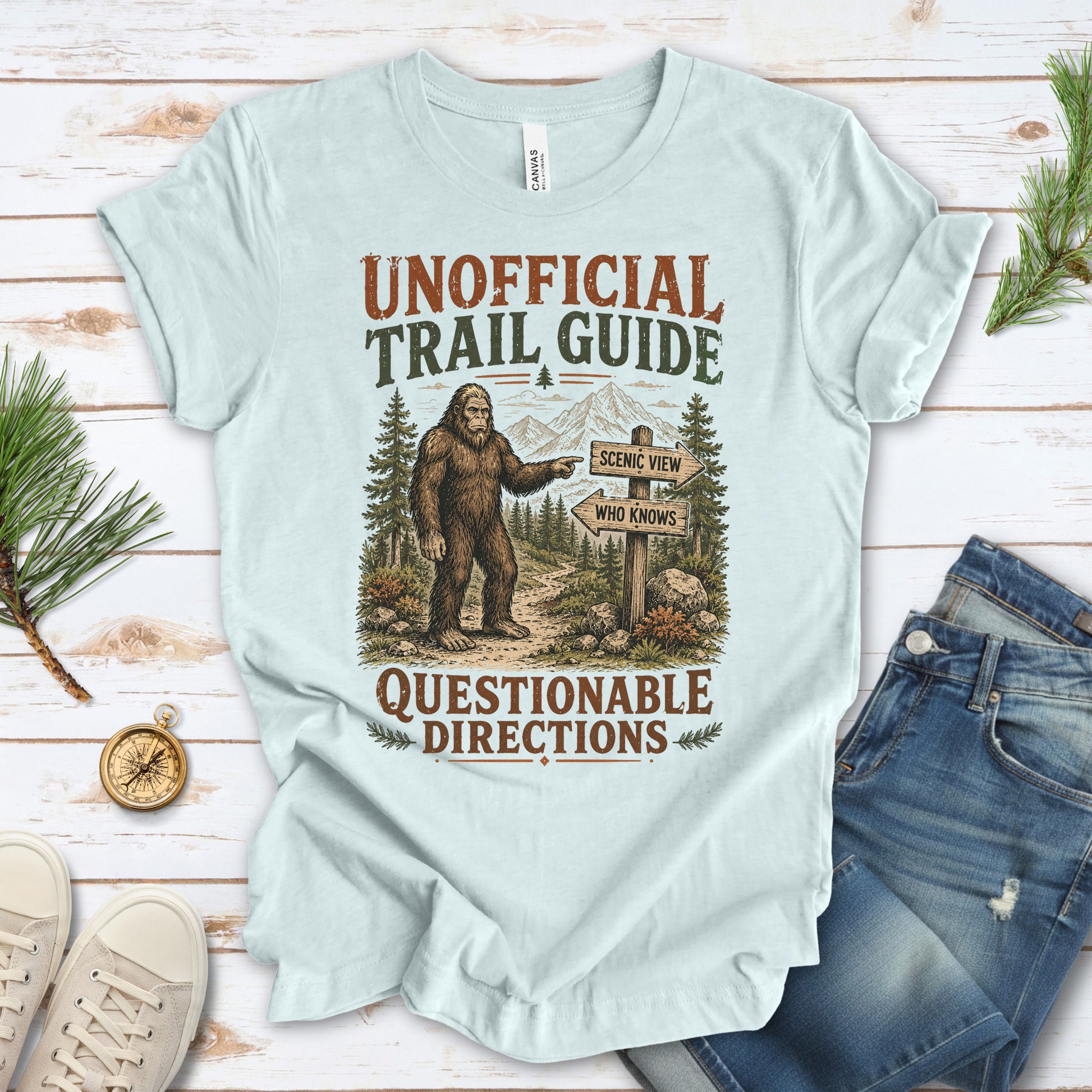 A light blue graphic t-shirt laid flat on a white wood background, styled with jeans, sneakers, pine branches, and a compass. The t-shirt features a vintage-style illustration of a Bigfoot figure pointing to road signs that read 'SCARY STUPID WHO KNOWS' and 'QUESTIONABLE DIRECTIONS'.