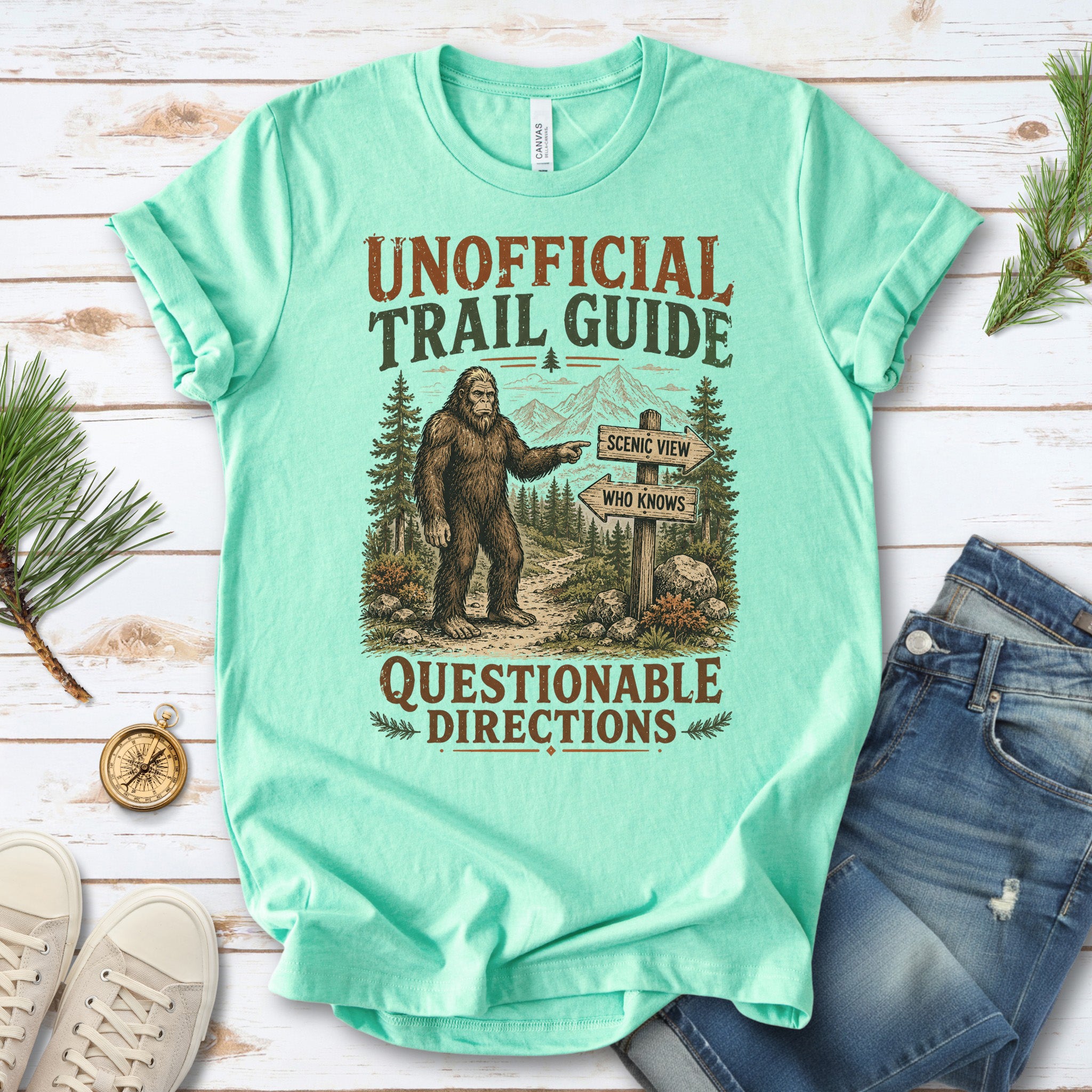 A mint green graphic t-shirt laid flat on a white wood background, styled with blue jeans, white sneakers, pine branches, and a small compass. The t-shirt features a graphic of Bigfoot pointing to a trail sign that reads 'SCENE VIEW WILD AFRICAN'.