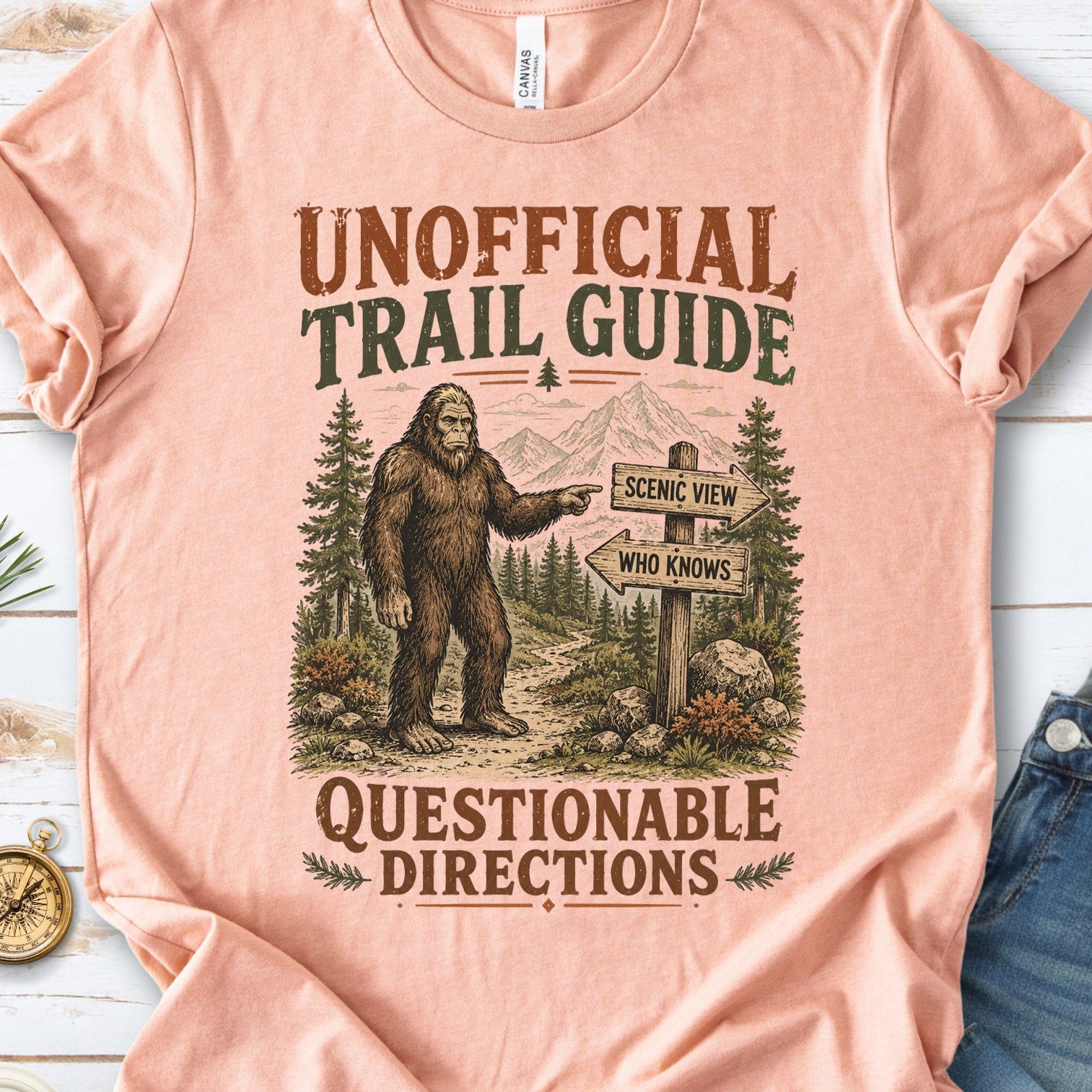 A peach-colored graphic t-shirt laid flat on a white wood background, styled with blue jeans, white sneakers, pine branches, and a small compass. The t-shirt features a vintage-style illustration of a Bigfoot figure pointing toward mountains and a trail signpost.