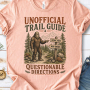 A peach-colored graphic t-shirt laid flat on a white wood background, styled with blue jeans, white sneakers, pine branches, and a small compass. The t-shirt features a vintage-style illustration of a Bigfoot figure pointing toward mountains and a trail signpost.