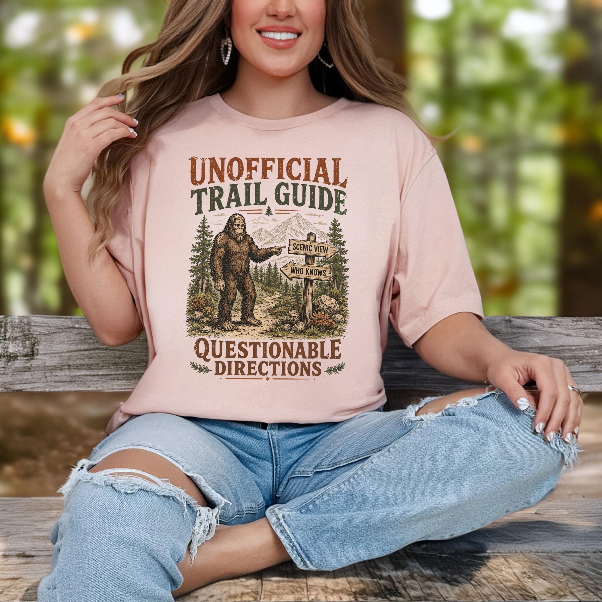 A woman sitting on a wooden bench outdoors wearing a light pink graphic t-shirt with a 'Unofficial Trail Guide' Bigfoot design and distressed light blue jeans.