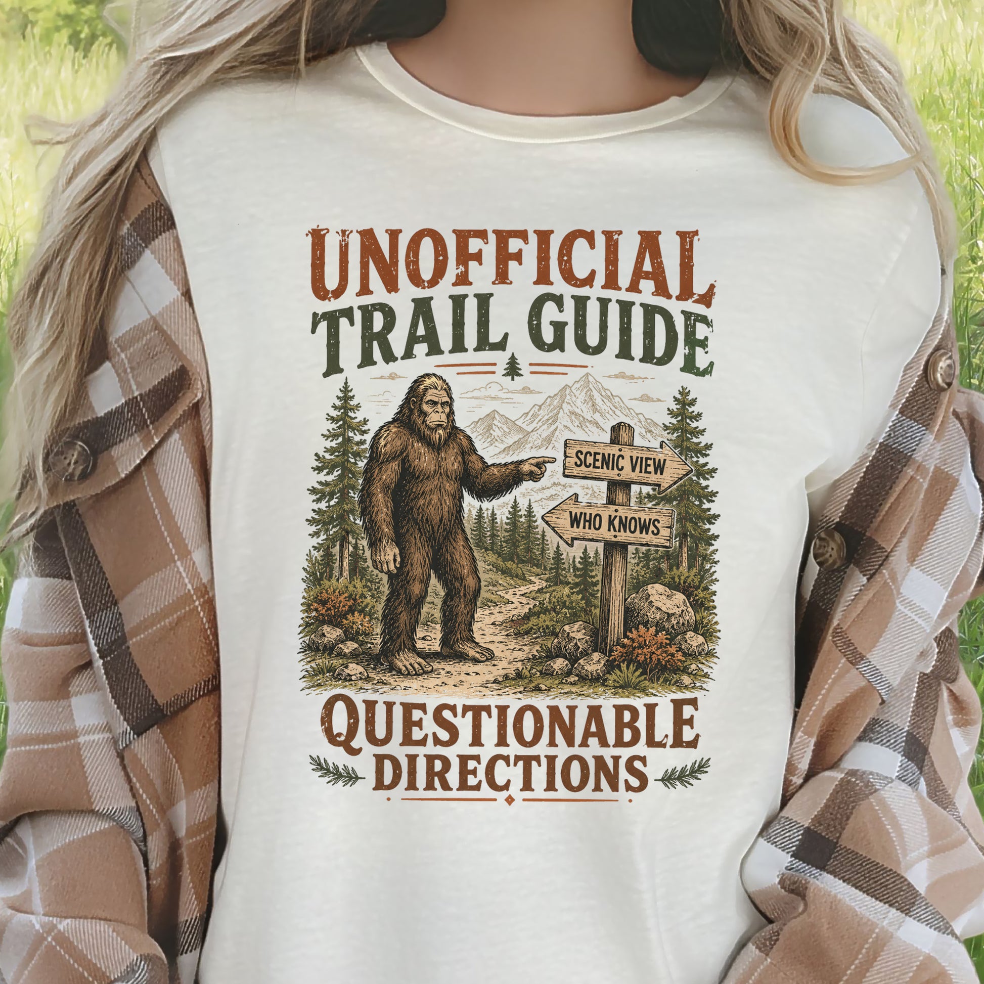 A person wearing a cream-colored graphic t-shirt with a brown and green 'Unofficial Trail Guide' Bigfoot design, layered with a brown and beige plaid flannel shirt, set against a blurred green outdoor background.
