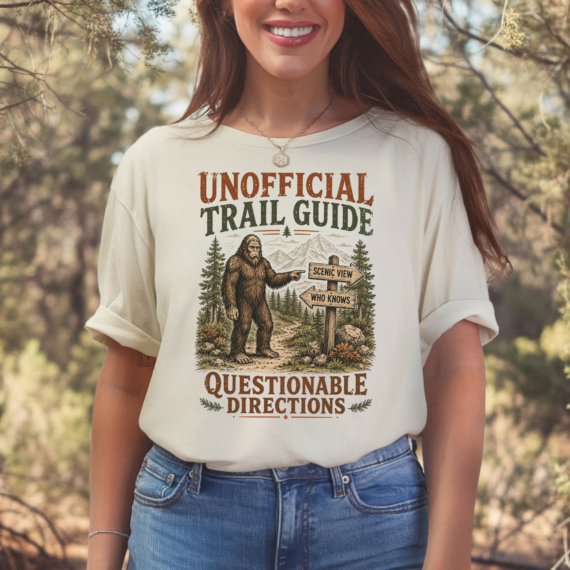 A woman wearing a cream-colored graphic t-shirt with a 'Bigfoot Trail Guide' design, standing outdoors in a blurred forest setting.