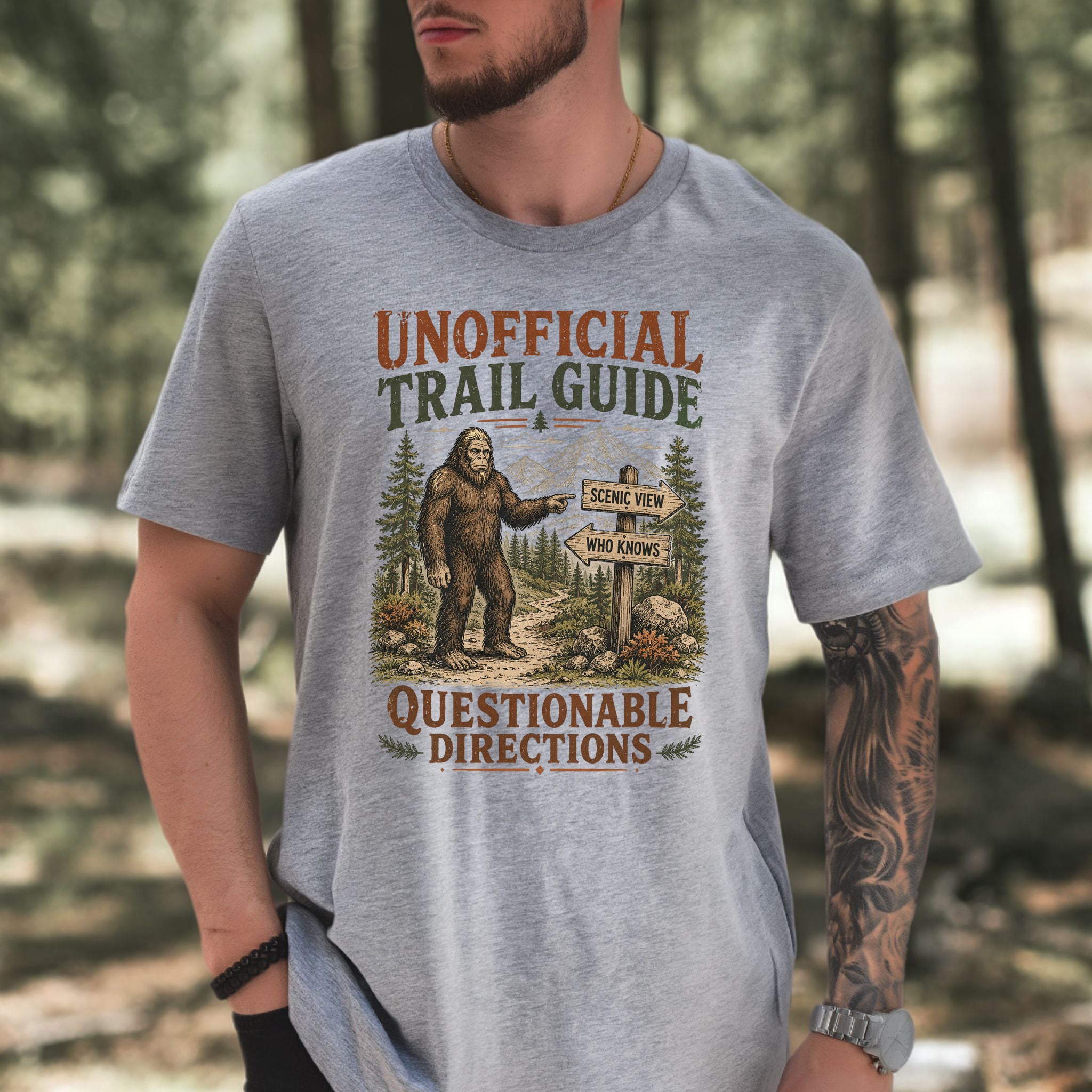 A man wearing a heather gray graphic t-shirt with a Bigfoot trail design, standing in a blurred forest setting.
