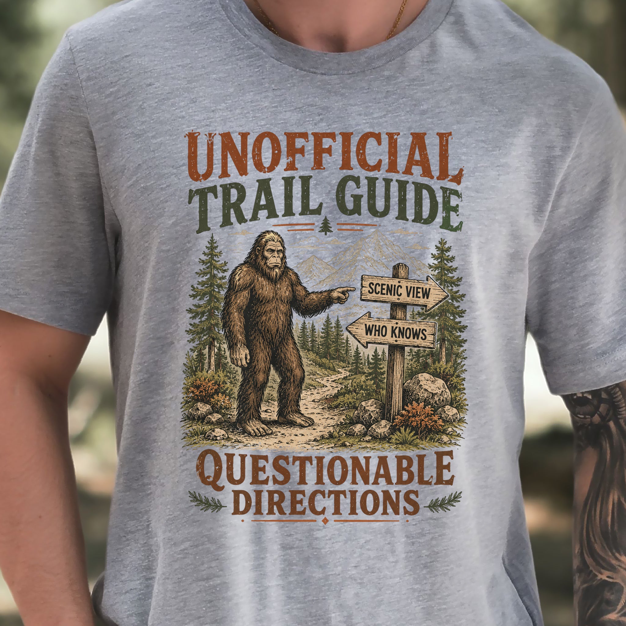 A close-up of a person wearing a heather gray t-shirt with a vintage-style graphic of Bigfoot pointing to a trail sign that reads 'SCENE VIEW WHO KNOWS' and 'QUESTIONABLE DIRECTIONS'.