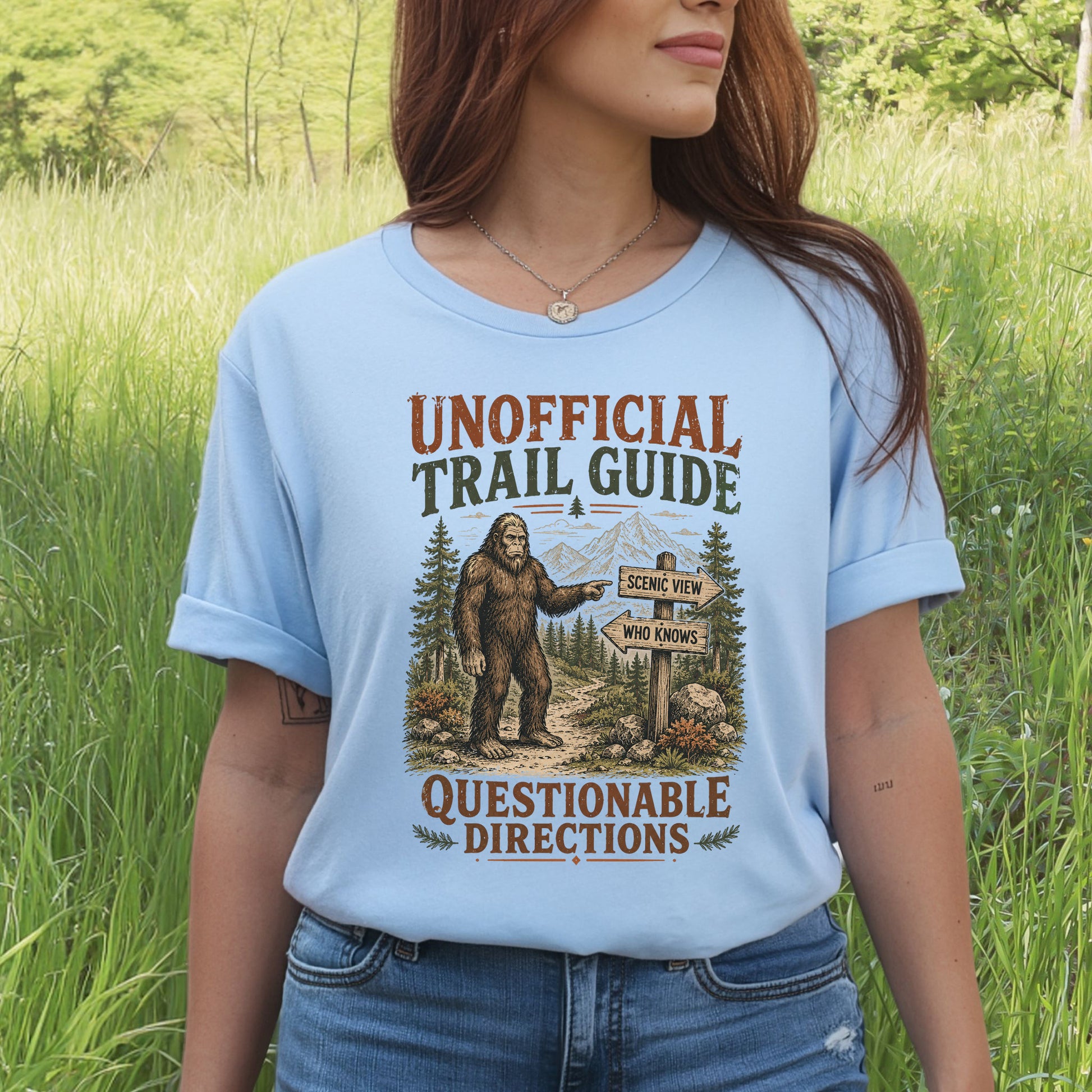 A woman wearing a light blue graphic t-shirt in an outdoor grassy field. The t-shirt features a retro-style graphic with the text 'UNOFFICIAL TRAIL GUIDE' and 'QUESTIONABLE DIRECTIONS' surrounding an illustration of Bigfoot pointing to a trail sign.