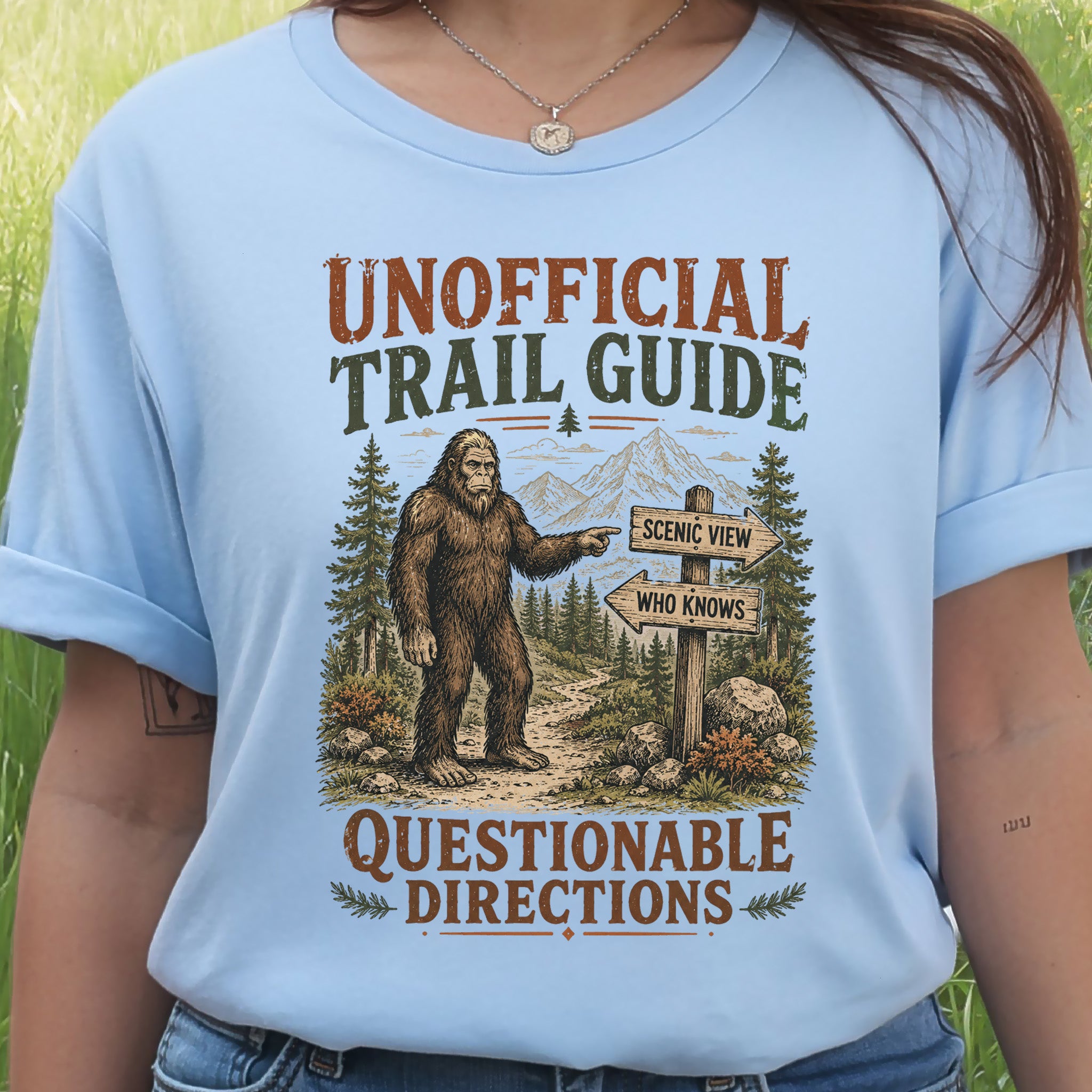 A person wearing a light blue graphic T-shirt outdoors. The shirt features a large front print with the words 'UNOFFICIAL TRAIL GUIDE' and 'QUESTIONABLE DIRECTIONS,' plus an illustration of Bigfoot pointing toward mountains and a trail sign that says 'SCENIC VIEW' and 'WHO KNOWS.