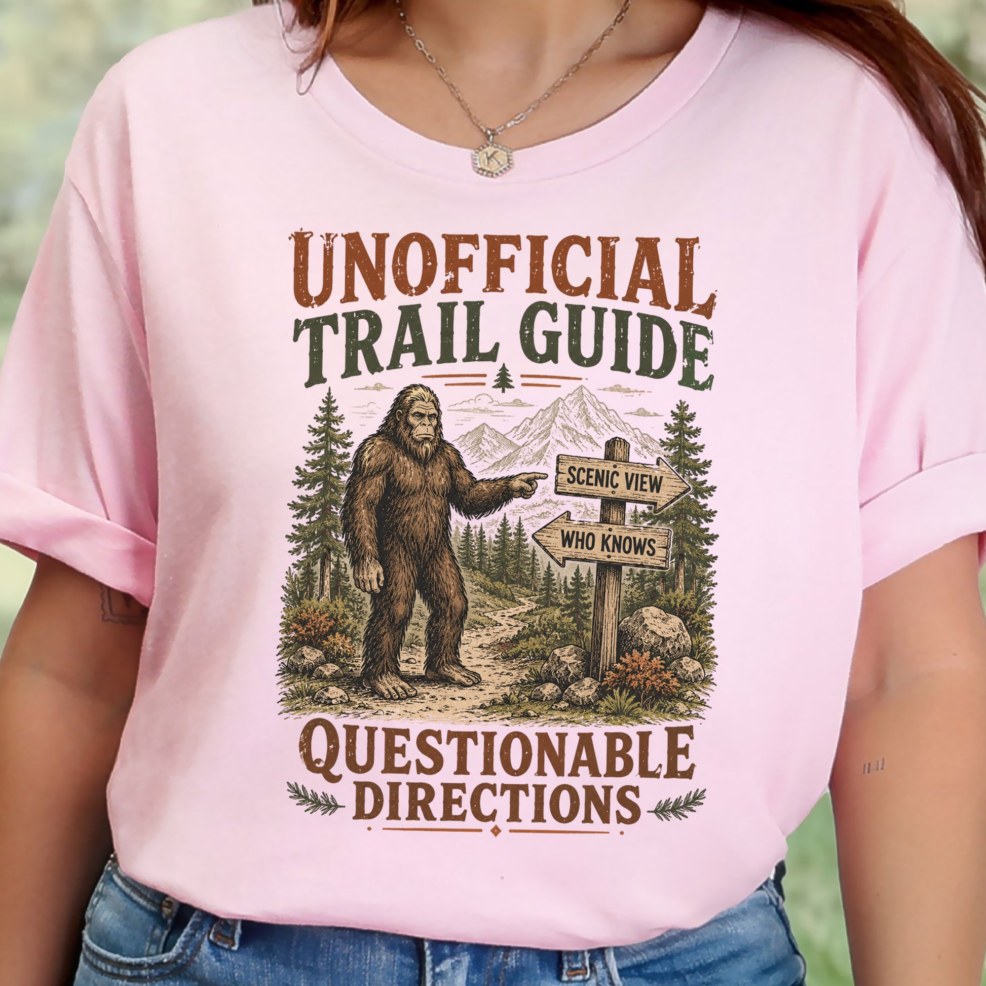 A woman wearing a light pink graphic t-shirt with a vintage-style print of Bigfoot and trail signs reading 'UNOFFICIAL TRAIL GUIDE' and 'QUESTIONABLE DIRECTIONS'.