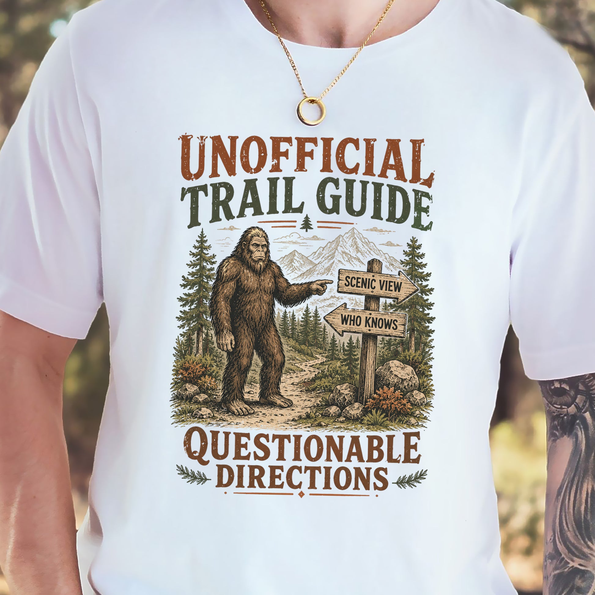 A white short-sleeve graphic t-shirt worn by a person, featuring a cartoon Bigfoot pointing to a trail sign that says 'SCENIC VIEW' and 'WHO KNOWS', with the text 'UNOFFICIAL TRAIL GUIDE' above and 'QUESTIONABLE DIRECTIONS' below.