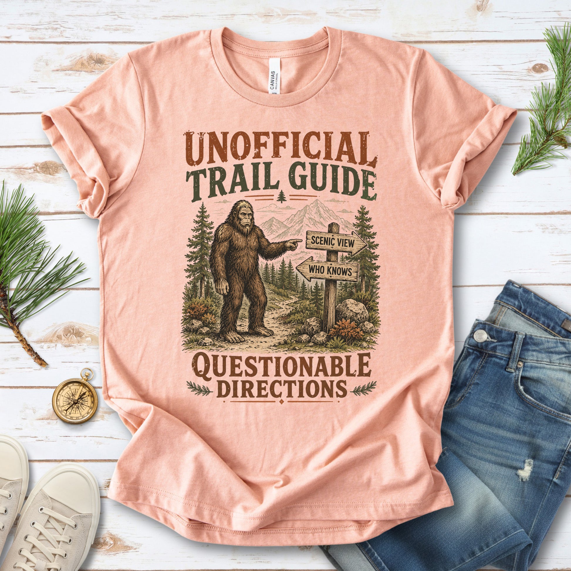 A peach-colored graphic t-shirt laid flat on a white wood background, styled with blue jeans, white sneakers, pine branches, and a small compass. The t-shirt features a vintage-style illustration of a Bigfoot figure pointing toward mountains and a trail signpost.